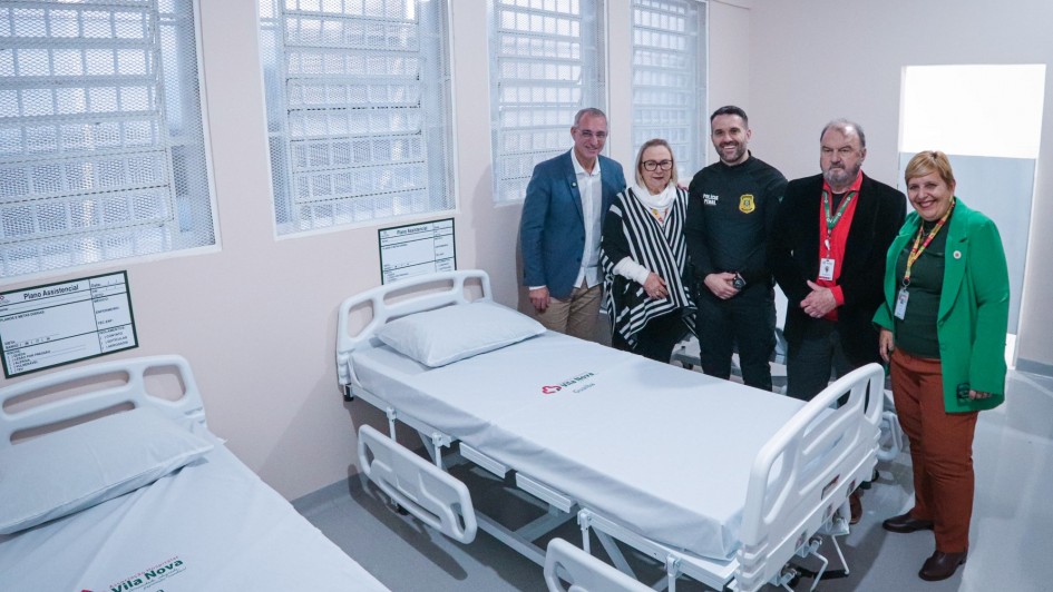 Esta é uma fotografia tirada em um quarto de hospital recém-inaugurado, mostrando camas hospitalares e um grupo de cinco pessoas adultas posando.
No primeiro plano, há duas camas hospitalares de metal branco com colchões claros e roupas de cama brancas. A cama em primeiro plano à esquerda tem a inscrição "Vila Nova Guaíba" bordada na roupa de cama. Nas paredes claras do quarto, ao lado de cada cama, há quadros brancos com formulários ou informações médicas.
Ao fundo, perto de uma grande janela com grades e persianas horizontais, um grupo de cinco pessoas está em pé e posando para a foto. Da esquerda para a direita:
Homem de blazer azul: Veste um blazer azul-claro sobre uma camisa branca e calça cáqui.
Mulher de óculos e echarpe: Veste óculos, uma blusa escura e uma echarpe listrada em preto e branco.
Homem de uniforme escuro: Veste um uniforme escuro com distintivos no ombro, semelhante a um guarda ou oficial.
Homem de blazer preto: Veste um blazer preto, camisa vermelha e gravata preta. Ele tem um crachá de identificação pendurado no pescoço.
Mulher de blazer verde: Veste um blazer verde-esmeralda sobre uma blusa escura e calças marrons.
O ambiente é limpo e bem iluminado, sugerindo um espaço funcional e recém-preparado.
