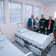 Esta é uma fotografia tirada em um quarto de hospital recém-inaugurado, mostrando camas hospitalares e um grupo de cinco pessoas adultas posando.

No primeiro plano, há duas camas hospitalares de metal branco com colchões claros e roupas de cama brancas. A cama em primeiro plano à esquerda tem a inscrição "Vila Nova Guaíba" bordada na roupa de cama. Nas paredes claras do quarto, ao lado de cada cama, há quadros brancos com formulários ou informações médicas.

Ao fundo, perto de uma grande janela com grades e persianas horizontais, um grupo de cinco pessoas está em pé e posando para a foto. Da esquerda para a direita:

Homem de blazer azul: Veste um blazer azul-claro sobre uma camisa branca e calça cáqui.

Mulher de óculos e echarpe: Veste óculos, uma blusa escura e uma echarpe listrada em preto e branco.

Homem de uniforme escuro: Veste um uniforme escuro com distintivos no ombro, semelhante a um guarda ou oficial.

Homem de blazer preto: Veste um blazer preto, camisa vermelha e gravata preta. Ele tem um crachá de identificação pendurado no pescoço.

Mulher de blazer verde: Veste um blazer verde-esmeralda sobre uma blusa escura e calças marrons.

O ambiente é limpo e bem iluminado, sugerindo um espaço funcional e recém-preparado.