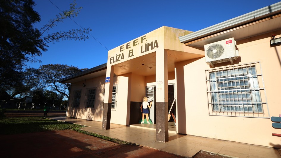 A imagem mostra a entrada principal de uma escola em um dia ensolarado. A fachada do prédio é bege e marrom escuro na parte inferior. Acima da porta de entrada, em letras grandes e escuras, lê-se "E. E. E. F. ELIZA B. LIMA". À esquerda da porta, há um adesivo vermelho e azul com a palavra "ROTA" e a frase "Segurança em Primeiro Lugar". No canto superior direito da imagem, um aparelho de ar condicionado está fixado na parede externa. Através da porta aberta, é possível ver um interior iluminado e algumas figuras de papelão, incluindo uma representação de uma menina e um cavalete com um desenho colorido.