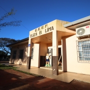 A imagem mostra a entrada principal de uma escola em um dia ensolarado. A fachada do prédio é bege e marrom escuro na parte inferior. Acima da porta de entrada, em letras grandes e escuras, lê-se "E. E. E. F. ELIZA B. LIMA". À esquerda da porta, há um adesivo vermelho e azul com a palavra "ROTA" e a frase "Segurança em Primeiro Lugar". No canto superior direito da imagem, um aparelho de ar condicionado está fixado na parede externa. Através da porta aberta, é possível ver um interior iluminado e algumas figuras de papelão, incluindo uma representação de uma menina e um cavalete com um desenho colorido.