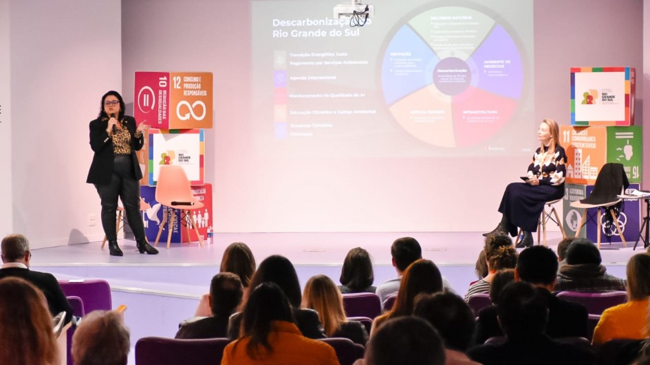 A imagem retrata uma apresentação em um auditório, com duas mulheres no palco e uma plateia sentada de costas para o observador.
No centro do palco, à esquerda, uma mulher de óculos, blusa com estampa de onça e calças escuras está em pé, segurando um microfone e gesticulando enquanto fala. Atrás dela, há um totem com o logotipo dos 17 Objetivos de Desenvolvimento Sustentável (ODS) da ONU, destacando os objetivos "10 Redução das Desigualdades" e "12 Consumo e Produção Responsáveis".
À direita, sentada em uma cadeira, está outra mulher, vestindo uma blusa estampada de flores e uma saia escura.
No fundo do palco, um grande telão exibe um infográfico circular colorido, com o título "Descarbonização Rio Grande do Sul". O gráfico apresenta seções como "Inovação", "Ambiente de Negócios", "Descarbonização", "Infraestrutura", "Gestão Urbana", "Governo Climático" e "Educação". Além disso, o telão lista tópicos como "Transição Energética Justa", "Pagamento por Serviços Ambientais", "Agenda Internacional", "Monitoramento da Qualidade do Ar", "Educação Climática e Justiça Ambiental", "Governança Climática" e "Inovação".
À direita do telão, há um banner com o título "11º Fórum Gaúcho de Mudanças Climáticas", acompanhado de outros logotipos relacionados aos ODS. A plateia ocupa as primeiras filas do auditório, com cadeiras roxas, e a maioria das pessoas está de costas, prestando atenção à apresentação.