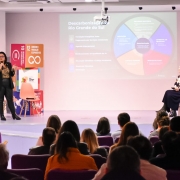 A imagem retrata uma apresentação em um auditório, com duas mulheres no palco e uma plateia sentada de costas para o observador.

No centro do palco, à esquerda, uma mulher de óculos, blusa com estampa de onça e calças escuras está em pé, segurando um microfone e gesticulando enquanto fala. Atrás dela, há um totem com o logotipo dos 17 Objetivos de Desenvolvimento Sustentável (ODS) da ONU, destacando os objetivos "10 Redução das Desigualdades" e "12 Consumo e Produção Responsáveis".

À direita, sentada em uma cadeira, está outra mulher, vestindo uma blusa estampada de flores e uma saia escura.

No fundo do palco, um grande telão exibe um infográfico circular colorido, com o título "Descarbonização Rio Grande do Sul". O gráfico apresenta seções como "Inovação", "Ambiente de Negócios", "Descarbonização", "Infraestrutura", "Gestão Urbana", "Governo Climático" e "Educação". Além disso, o telão lista tópicos como "Transição Energética Justa", "Pagamento por Serviços Ambientais", "Agenda Internacional", "Monitoramento da Qualidade do Ar", "Educação Climática e Justiça Ambiental", "Governança Climática" e "Inovação".

À direita do telão, há um banner com o título "11º Fórum Gaúcho de Mudanças Climáticas", acompanhado de outros logotipos relacionados aos ODS. A plateia ocupa as primeiras filas do auditório, com cadeiras roxas, e a maioria das pessoas está de costas, prestando atenção à apresentação.