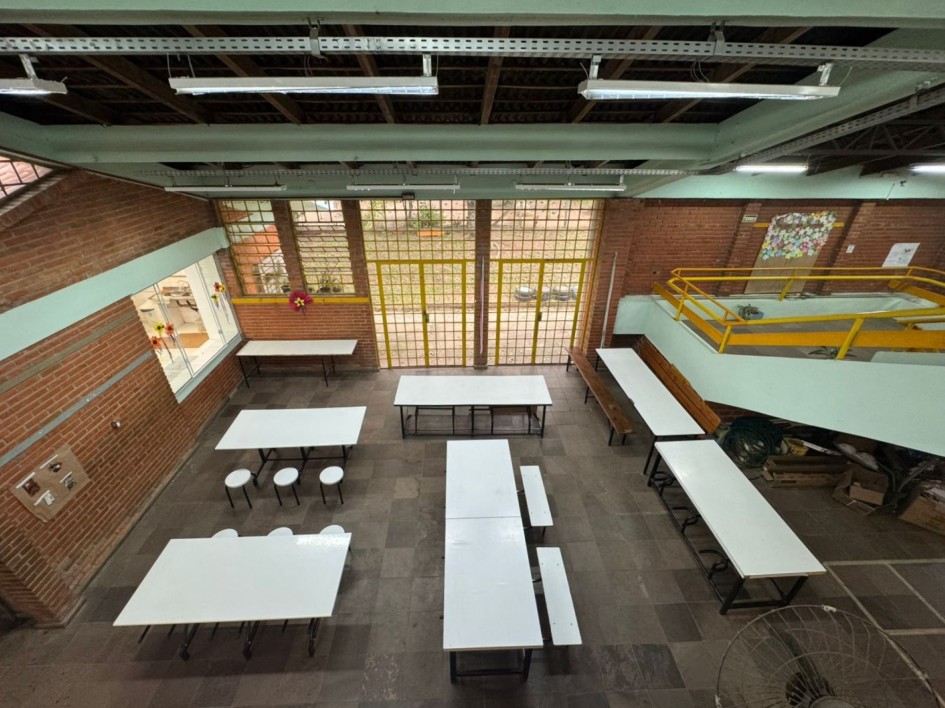 Espaço amplo com mesas e bancos brancos, semelhante a um refeitório escolar. À esquerda, uma janela com flores decorativas traz luz ao ambiente. À frente, parede de tijolos com portas de vidro que levam para fora. Acima, há um mezanino com corrimão amarelo.