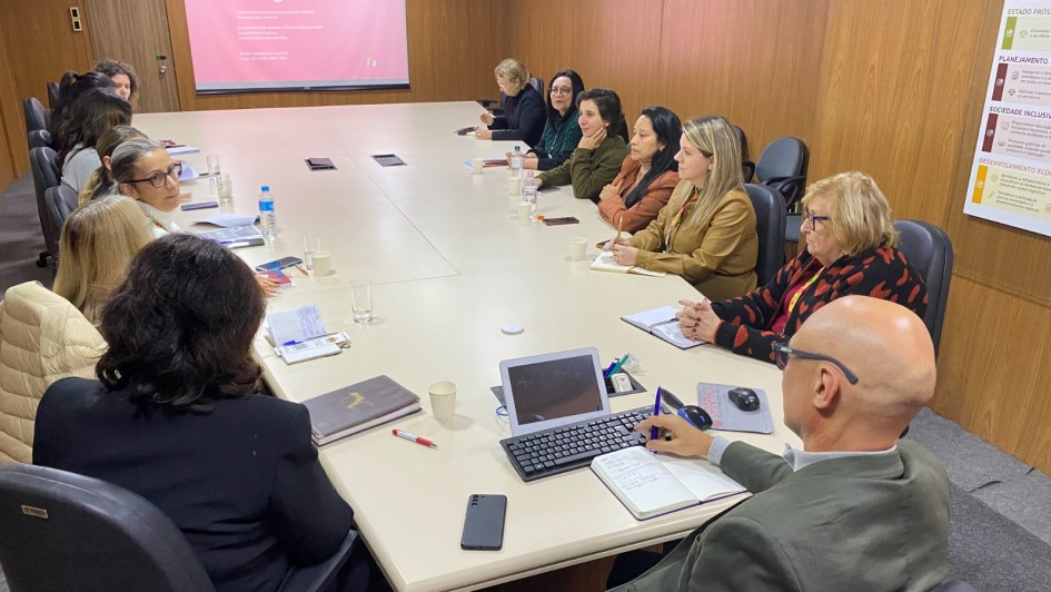 A imagem mostra uma sala de reunião com um grupo de pessoas sentadas ao redor de uma grande mesa retangular, que é bege clara. A maioria das pessoas são mulheres, com idades variadas, e todas parecem atentas, algumas olhando para o centro da mesa ou para a tela de projeção.

No primeiro plano, de costas para o observador, um homem calvo com óculos veste uma jaqueta verde oliva e está usando um notebook e anotando algo em um caderno. Ao lado dele, há um smartphone preto.

No fundo da sala, um projetor está ligado e exibe a palavra "Obrigada!" em letras brancas sobre um fundo rosa claro na parede. Há também um banner vertical parcialmente visível à direita, com a palavra "MAPA" no topo.

A mesa está repleta de papéis, cadernos, copos com água e alguns laptops, indicando uma reunião de trabalho. As cadeiras são escuras, com braços, e as paredes da sala são de um tom claro, com painéis de madeira.