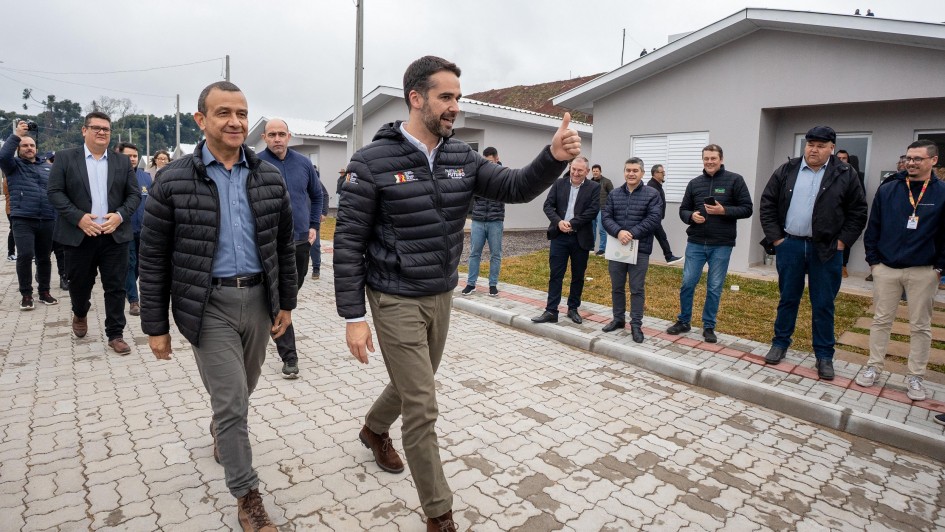 A foto mostra o Governador Eduardo Leite, à direita, com um casaco puffer preto e calças cáqui, caminhando e fazendo um sinal de "joinha" com o polegar para cima. Ao seu lado esquerdo, o Secretário Carlos Gomes, com um casaco puffer escuro e calças cinzas, caminha olhando para frente. Ambos sorriem enquanto andam sobre um calçamento de blocos intertravados em Santo Antônio do Palma. Ao fundo, diversas casas cinzas recém-construídas são visíveis, indicando a entrega de moradias. Um grupo de homens, alguns com celulares, acompanha e observa a caminhada dos dois políticos, completando a cena da inauguração.