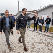 A foto mostra o Governador Eduardo Leite, à direita, com um casaco puffer preto e calças cáqui, caminhando e fazendo um sinal de "joinha" com o polegar para cima. Ao seu lado esquerdo, o Secretário Carlos Gomes, com um casaco puffer escuro e calças cinzas, caminha olhando para frente. Ambos sorriem enquanto andam sobre um calçamento de blocos intertravados em Santo Antônio do Palma. Ao fundo, diversas casas cinzas recém-construídas são visíveis, indicando a entrega de moradias. Um grupo de homens, alguns com celulares, acompanha e observa a caminhada dos dois políticos, completando a cena da inauguração.