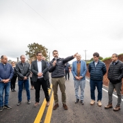 A foto mostra o Governador Eduardo Leite, no centro, falando em um microfone enquanto gesticula com a mão direita, inaugurando uma obra de pavimentação asfáltica em Santo Antônio do Palma. Ele veste um casaco puffer preto e calças cáqui. Ao seu redor, um grupo de aproximadamente dez homens o ouve atentamente, a maioria vestindo casacos e calças jeans ou sociais. A cena se passa em uma estrada recém-pavimentada, marcada com faixas amarelas duplas no asfalto. À direita da imagem, um talude de terra avermelhada indica a lateral da estrada, e ao fundo, o céu está nublado, com árvores e vegetação rasteira em ambos os lados da via.