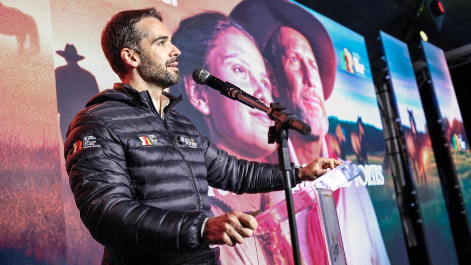A foto mostra o Governador Eduardo Leite em pé, falando em um microfone durante o lançamento da 48ª Expointer. Ele veste um casaco puffer preto e segura uma pasta transparente sobre um púlpito também transparente. O governador olha para a direita, com uma expressão séria e concentrada. Ao fundo, uma grande tela exibe uma imagem artística em tons sépia, com rostos de pessoas e silhuetas de cavalos no campo, remetendo ao tema gaúcho e rural da Expointer. O ambiente é iluminado por luzes de palco, e o fundo do palco é composto por várias telas alinhadas.