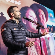 A foto mostra o Governador Eduardo Leite em pé, falando em um microfone durante o lançamento da 48ª Expointer. Ele veste um casaco puffer preto e segura uma pasta transparente sobre um púlpito também transparente. O governador olha para a direita, com uma expressão séria e concentrada. Ao fundo, uma grande tela exibe uma imagem artística em tons sépia, com rostos de pessoas e silhuetas de cavalos no campo, remetendo ao tema gaúcho e rural da Expointer. O ambiente é iluminado por luzes de palco, e o fundo do palco é composto por várias telas alinhadas.