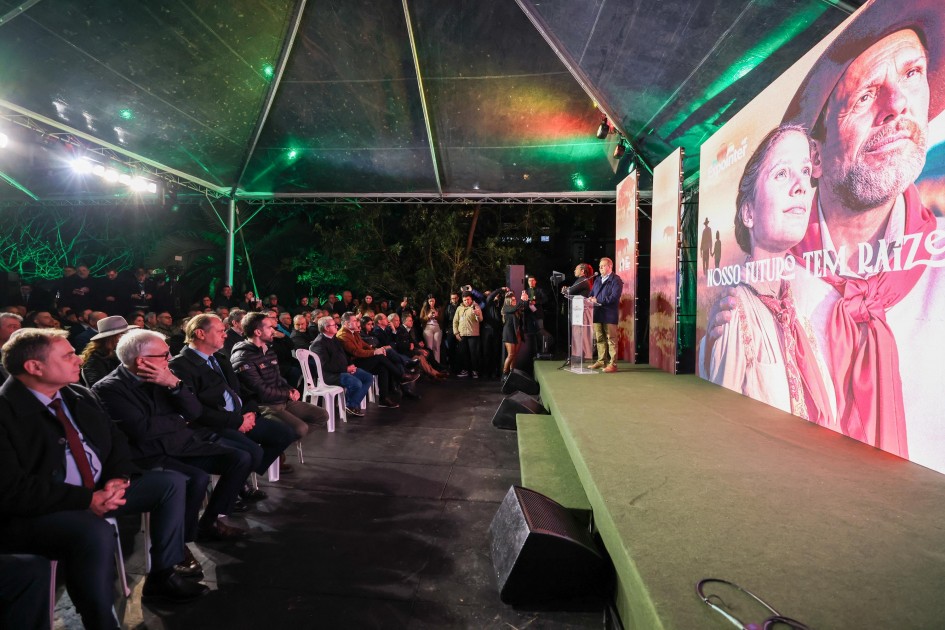 A foto mostra o ambiente de lançamento da 48ª Expointer, com um palco iluminado à direita e uma plateia sentada à esquerda, sob uma tenda translúcida. No palco, um homem fala ao microfone em um púlpito, com grandes telas atrás dele exibindo imagens artísticas de rostos de gaúchos e a frase "NOSSO FUTURO TEM RAÍZES". A plateia, composta por diversas pessoas, majoritariamente homens, está sentada em cadeiras brancas e observa o palco. O local está sob uma iluminação verde vibrante, que cria uma atmosfera de evento noturno.