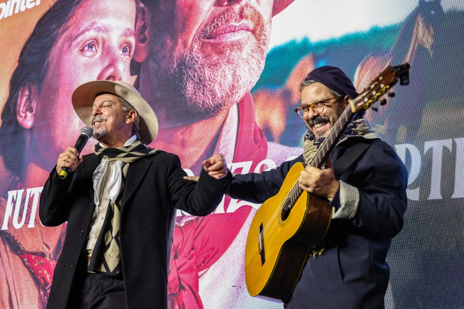 A foto mostra dois homens em um palco, com grandes telas ao fundo exibindo imagens artísticas de rostos e a palavra "FUTURO". À esquerda, o cantor Neto Fagundes, com chapéu de aba larga, lenço no pescoço e casaco escuro, canta em um microfone, com o braço esquerdo estendido em um gesto de interação. À direita, um músico sorri, usando gorro, óculos e um lenço no pescoço, enquanto toca um violão clássico, também com o braço direito estendido. A cena transmite a alegria e a energia de uma apresentação musical durante o evento de lançamento da Expointer.