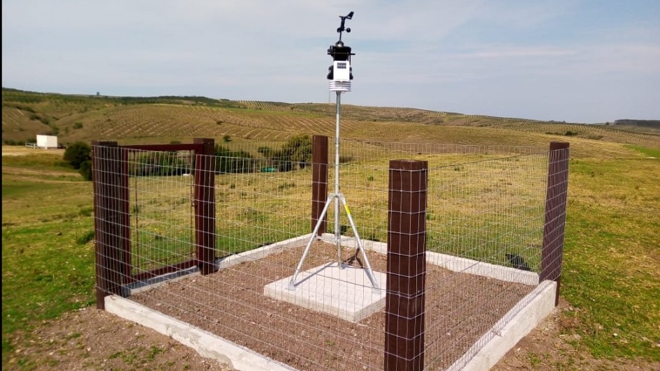 A imagem mostra uma estação meteorológica automática instalada em um campo aberto, cercada por uma estrutura de proteção com tela de arame e postes de madeira. A estação está montada sobre um tripé metálico no centro da área cercada, com sensores na parte superior para medir dados como temperatura, vento e umidade. Ao fundo, há colinas verdes e céu azul com algumas nuvens finas.