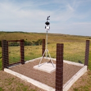 A imagem mostra uma estação meteorológica automática instalada em um campo aberto, cercada por uma estrutura de proteção com tela de arame e postes de madeira. A estação está montada sobre um tripé metálico no centro da área cercada, com sensores na parte superior para medir dados como temperatura, vento e umidade. Ao fundo, há colinas verdes e céu azul com algumas nuvens finas.