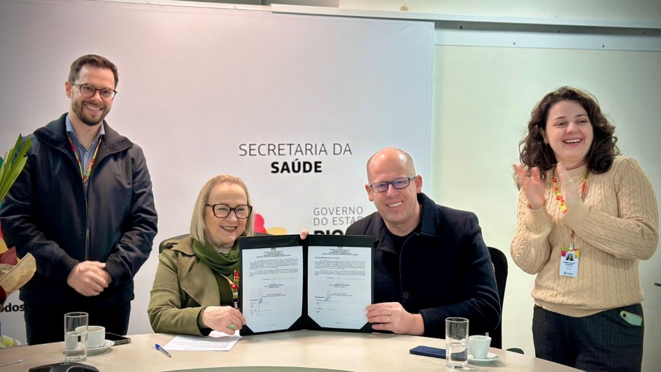 Esta é uma fotografia colorida de quatro pessoas, dois homens e duas mulheres, em um ambiente de escritório ou sala de reuniões, celebrando a assinatura de um documento.

No centro da imagem, sentados a uma mesa curva de cor clara, uma mulher de óculos com cabelos curtos e claros, vestindo um casaco verde-militar e uma echarpe verde, e um homem careca de óculos, vestindo um casaco escuro, sorriem enquanto seguram juntos um documento aberto com assinaturas.

Atrás deles, há uma parede branca com a inscrição "SECRETARIA DA SAÚDE" em letras escuras, e abaixo, "GOVERNO DO ESTADO".

À esquerda do casal central, um homem com barba e óculos, vestindo uma jaqueta escura sobre uma camisa quadriculada, está em pé e sorri para a câmera. À direita do casal, uma mulher com cabelos cacheados e compridos, vestindo um suéter bege, está em pé e aplaudindo, com um sorriso.

Sobre a mesa, há copos com água, xícaras de café e alguns objetos. A iluminação é clara e o ambiente parece ser formal.
