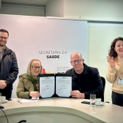 Esta é uma fotografia colorida de quatro pessoas, dois homens e duas mulheres, em um ambiente de escritório ou sala de reuniões, celebrando a assinatura de um documento.

No centro da imagem, sentados a uma mesa curva de cor clara, uma mulher de óculos com cabelos curtos e claros, vestindo um casaco verde-militar e uma echarpe verde, e um homem careca de óculos, vestindo um casaco escuro, sorriem enquanto seguram juntos um documento aberto com assinaturas.

Atrás deles, há uma parede branca com a inscrição "SECRETARIA DA SAÚDE" em letras escuras, e abaixo, "GOVERNO DO ESTADO".

À esquerda do casal central, um homem com barba e óculos, vestindo uma jaqueta escura sobre uma camisa quadriculada, está em pé e sorri para a câmera. À direita do casal, uma mulher com cabelos cacheados e compridos, vestindo um suéter bege, está em pé e aplaudindo, com um sorriso.

Sobre a mesa, há copos com água, xícaras de café e alguns objetos. A iluminação é clara e o ambiente parece ser formal.