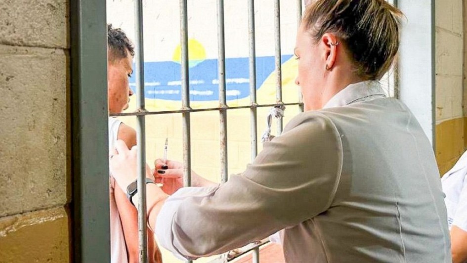 A foto mostra uma cena de vacinação em um ambiente prisional. Uma mulher, provavelmente uma profissional de saúde, com cabelo preso em coque, veste uma camisa clara e está de perfil, à direita. Ela aplica uma injeção no braço esquerdo de um homem que aparece à esquerda, atrás das grades verticais de uma cela. O homem, com pele escura e cabelo crespo, está de perfil, olhando para frente. Ao fundo, através das grades, é visível uma parede pintada com um desenho que simula um céu azul, um sol amarelo e montanhas, ou algo semelhante a uma paisagem marinha, adicionando um contraste de cores vibrantes ao ambiente austero. As grades e as paredes de pedra rústica dominam o primeiro plano e os lados da imagem, reforçando a ideia de confinamento.