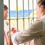 A foto mostra uma cena de vacinação em um ambiente prisional. Uma mulher, provavelmente uma profissional de saúde, com cabelo preso em coque, veste uma camisa clara e está de perfil, à direita. Ela aplica uma injeção no braço esquerdo de um homem que aparece à esquerda, atrás das grades verticais de uma cela. O homem, com pele escura e cabelo crespo, está de perfil, olhando para frente. Ao fundo, através das grades, é visível uma parede pintada com um desenho que simula um céu azul, um sol amarelo e montanhas, ou algo semelhante a uma paisagem marinha, adicionando um contraste de cores vibrantes ao ambiente austero. As grades e as paredes de pedra rústica dominam o primeiro plano e os lados da imagem, reforçando a ideia de confinamento.