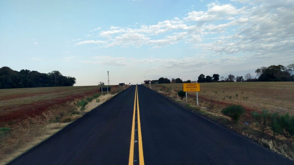 Esta é uma fotografia de uma estrada asfaltada reta que se estende até o horizonte sob um céu com nuvens esparsas.

A estrada, de cor preta escura, tem duas linhas amarelas contínuas no centro, indicando duas vias. Ela se projeta para o fundo da imagem, parecendo se afunilar no horizonte.

Nas laterais da estrada, há faixas de terra avermelhada e campos verdes com algumas árvores e vegetação rasteira. À direita, um pouco mais adiante na estrada, há uma placa de trânsito retangular amarela com a inscrição em letras pretas: "ATENÇÃO PEDESTRES A 200 M". Postes de eletricidade e algumas árvores distantes são visíveis ao longo da estrada. O céu está predominantemente claro, com algumas nuvens brancas e finas.