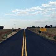 Esta é uma fotografia de uma estrada asfaltada reta que se estende até o horizonte sob um céu com nuvens esparsas.

A estrada, de cor preta escura, tem duas linhas amarelas contínuas no centro, indicando duas vias. Ela se projeta para o fundo da imagem, parecendo se afunilar no horizonte.

Nas laterais da estrada, há faixas de terra avermelhada e campos verdes com algumas árvores e vegetação rasteira. À direita, um pouco mais adiante na estrada, há uma placa de trânsito retangular amarela com a inscrição em letras pretas: "ATENÇÃO PEDESTRES A 200 M". Postes de eletricidade e algumas árvores distantes são visíveis ao longo da estrada. O céu está predominantemente claro, com algumas nuvens brancas e finas.