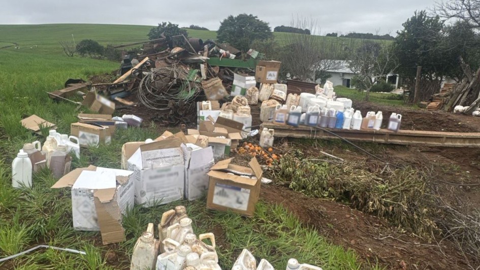 A imagem mostra uma área rural com muitos galões e caixas de agrotóxicos espalhados no chão, alguns sujos de terra e aparentemente usados. Ao fundo, há restos de materiais empilhados, como sucata e madeira. O cenário indica uma apreensão ou descarte irregular. A vegetação ao redor é verde, com um campo aberto e uma casa rural mais distante. O céu está nublado.