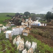 A imagem mostra uma área rural com muitos galões e caixas de agrotóxicos espalhados no chão, alguns sujos de terra e aparentemente usados. Ao fundo, há restos de materiais empilhados, como sucata e madeira. O cenário indica uma apreensão ou descarte irregular. A vegetação ao redor é verde, com um campo aberto e uma casa rural mais distante. O céu está nublado.