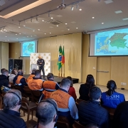 A foto mostra um seminário internacional da Defesa Civil ocorrendo em um auditório moderno. No centro do palco, um orador de costas para o observador, vestido com colete da Defesa Civil, fala para uma plateia atenta, composta em sua maioria por homens e mulheres também usando coletes laranjas ou uniformes escuros. Atrás do orador, em destaque, há duas bandeiras: uma do Brasil e outra do Rio Grande do Sul. Duas grandes telas de projeção, uma em cada lado do palco, exibem mapas coloridos da Europa, com destaque para a Itália, e outros dados relacionados. A iluminação é clara e uniforme, e o ambiente é organizado, com fileiras de cadeiras preenchidas. Alguns participantes usam fones de ouvido para tradução simultânea. A imagem sugere um evento de cooperação e aprendizado sobre temas de proteção civil.
