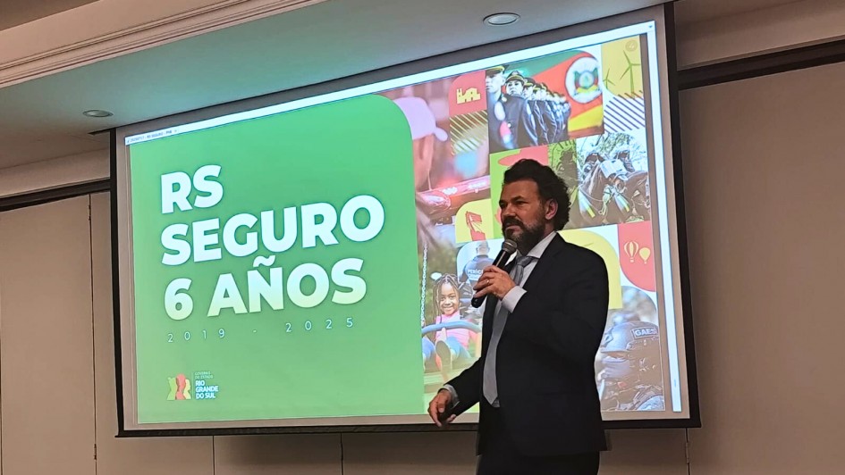 A foto mostra Antônio Padilha, Diretor Executivo do RS Seguro, um homem branco de cabelos e barba escura, vestindo terno e gravata, em pé no centro de um palco, segurando um microfone e gesticulando. Ao fundo, uma grande tela de projeção exibe a frase "RS SEGURO 6 ANOS" em letras grandes e brancas sobre um fundo verde vibrante. A tela também apresenta uma colagem de imagens relacionadas ao programa, incluindo cenas de policiamento e atividades comunitárias. O chão do palco tem um carpete estampado em tons de cinza. A iluminação geral do ambiente é clara e focada no orador e na projeção.