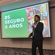 A foto mostra Antônio Padilha, Diretor Executivo do RS Seguro, um homem branco de cabelos e barba escura, vestindo terno e gravata, em pé no centro de um palco, segurando um microfone e gesticulando. Ao fundo, uma grande tela de projeção exibe a frase "RS SEGURO 6 ANOS" em letras grandes e brancas sobre um fundo verde vibrante. A tela também apresenta uma colagem de imagens relacionadas ao programa, incluindo cenas de policiamento e atividades comunitárias. O chão do palco tem um carpete estampado em tons de cinza. A iluminação geral do ambiente é clara e focada no orador e na projeção.