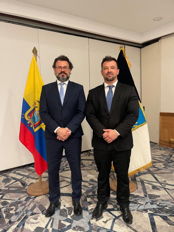 A foto mostra o Diretor Executivo do RS Seguro, Antônio Padilha, e o delegado Mario Souza em pé lado a lado, em um ambiente formal. Antônio Padilha, à esquerda, é um homem branco de barba e cabelo escuros, usando óculos, terno azul-marinho e gravata clara. O delegado Mario Souza, à direita, é um homem branco com barba e cabelo curtos, vestindo terno preto e gravata azul. Ambos têm as mãos unidas à frente do corpo. Atrás deles, há duas bandeiras hasteadas: uma do Equador, com suas faixas amarela, azul e vermelha e o brasão, e outra de cor preta e branca com detalhes em azul. O piso do local é um carpete estampado com padrões geométricos em tons de cinza e preto.