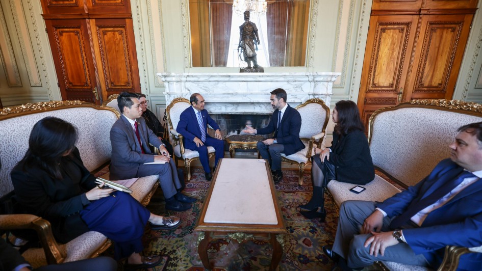 Uma reunião formal entre representantes do Estado e da Embaixada de Singapura acontece no Palácio Piratini. Sete pessoas vestindo trajes formais estão sentadas em poltronas clássicas com estofamento claro, dispostas em semicírculo ao redor de uma mesa de centro retangular de madeira clara e tampo de mármore. No centro da imagem, dois homens, um de terno azul e outro de terno escuro, estão engajados em uma conversa direta. Os demais participantes, incluindo duas mulheres, observam atentamente, e uma delas à esquerda segura um caderno. Ao fundo, o ambiente é luxuoso, com paredes de madeira escura, detalhes dourados, um lustre pendurado no teto e uma lareira ornamentada com uma estátua sobre ela.