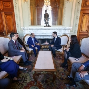Uma reunião formal entre representantes do Estado e da Embaixada de Singapura acontece no Palácio Piratini. Sete pessoas vestindo trajes formais estão sentadas em poltronas clássicas com estofamento claro, dispostas em semicírculo ao redor de uma mesa de centro retangular de madeira clara e tampo de mármore. No centro da imagem, dois homens, um de terno azul e outro de terno escuro, estão engajados em uma conversa direta. Os demais participantes, incluindo duas mulheres, observam atentamente, e uma delas à esquerda segura um caderno. Ao fundo, o ambiente é luxuoso, com paredes de madeira escura, detalhes dourados, um lustre pendurado no teto e uma lareira ornamentada com uma estátua sobre ela.