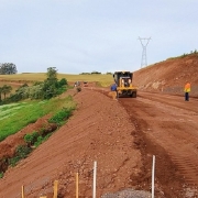 A imagem mostra uma estrada de chão em obras, com uma máquina ao centro e alguns trabalhadores uniformizados na pista. 
