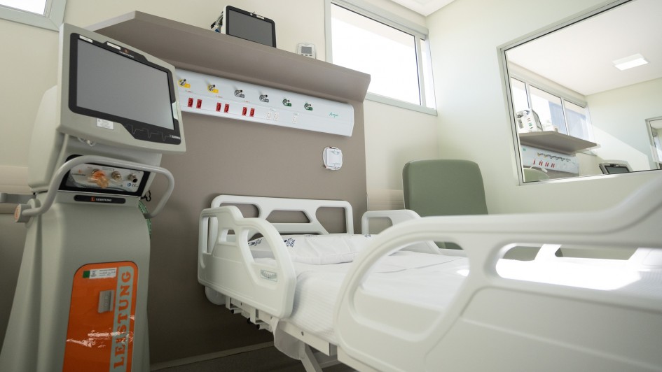 A foto mostra o interior de um quarto de hospital moderno, claro e bem equipado. No centro da imagem, uma cama hospitalar branca, vazia e com lençóis impecáveis, está posicionada. Acima da cabeceira da cama, na parede, há um painel com saídas para gases medicinais e tomadas elétricas. À esquerda da cama, um equipamento médico de grande porte, com uma tela e botões, sugere ser um monitor ou ventilador. Há janelas na parte superior da parede, permitindo a entrada de luz natural, e um espelho grande reflete parte do ambiente. Uma poltrona verde está posicionada ao lado direito da cama. O quarto transmite uma sensação de limpeza, organização e tecnologia.