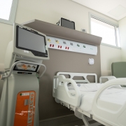A foto mostra o interior de um quarto de hospital moderno, claro e bem equipado. No centro da imagem, uma cama hospitalar branca, vazia e com lençóis impecáveis, está posicionada. Acima da cabeceira da cama, na parede, há um painel com saídas para gases medicinais e tomadas elétricas. À esquerda da cama, um equipamento médico de grande porte, com uma tela e botões, sugere ser um monitor ou ventilador. Há janelas na parte superior da parede, permitindo a entrada de luz natural, e um espelho grande reflete parte do ambiente. Uma poltrona verde está posicionada ao lado direito da cama. O quarto transmite uma sensação de limpeza, organização e tecnologia.
