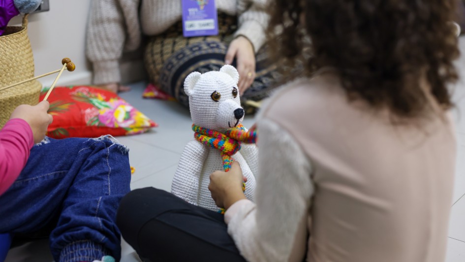 A imagem mostra uma criança de costas, com o cabelo cacheado. Ela está sentada no chão, brincando com um urso branco de cachecol colorido. Seu rosto não está visível. Ao lado, aparecem as pernas de outra criança sentada perto da menina e de uma pessoa adulta com crachá.