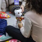 A imagem mostra uma criança de costas, com o cabelo cacheado. Ela está sentada no chão, brincando com um urso branco de cachecol colorido. Seu rosto não está visível. Ao lado, aparecem as pernas de outra criança sentada perto da menina e de uma pessoa adulta com crachá. 