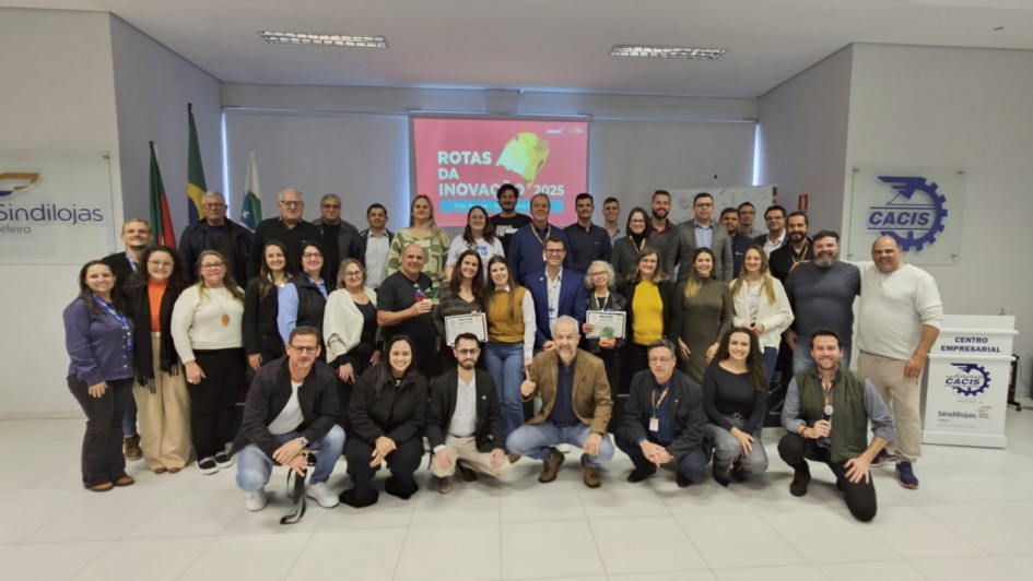 A foto mostra um grande grupo de aproximadamente quarenta pessoas, entre homens e mulheres de diversas idades, posando para uma foto de grupo. Eles estão em pé e agachados em um ambiente interno, que parece ser um auditório ou sala de eventos, com iluminação clara. Ao fundo, um telão projeta a frase "Rotas da Inovação 2025" em letras brancas sobre um fundo vermelho. Ao redor do telão, há banners e logotipos de várias empresas e organizações, como o Sindilojas e a Cacis. O grupo, em sua maioria, está sorrindo e olhando para a câmera, com alguns indivíduos segurando placas ou certificados.