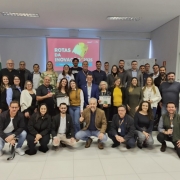 A foto mostra um grande grupo de aproximadamente quarenta pessoas, entre homens e mulheres de diversas idades, posando para uma foto de grupo. Eles estão em pé e agachados em um ambiente interno, que parece ser um auditório ou sala de eventos, com iluminação clara. Ao fundo, um telão projeta a frase "Rotas da Inovação 2025" em letras brancas sobre um fundo vermelho. Ao redor do telão, há banners e logotipos de várias empresas e organizações, como o Sindilojas e a Cacis. O grupo, em sua maioria, está sorrindo e olhando para a câmera, com alguns indivíduos segurando placas ou certificados.