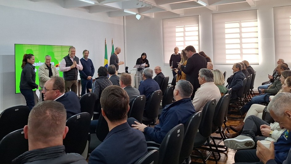 Uma foto mostra uma reunião em ambiente fechado com muitas pessoas, algumas em pé e outras sentadas. À esquerda, vários homens estão de pé perto de uma grande tela verde brilhante com algum texto visível. Uma mulher está à extrema esquerda deles, olhando para eles. No centro e à direita, uma plateia está sentada em fileiras de cadeiras escuras, de frente para a parte da frente onde estão os indivíduos em pé. Alguns membros da plateia estão olhando para a frente, enquanto outros estão olhando para seus telefones ou conversando entre si. A sala tem paredes de cores claras e janelas com persianas são visíveis no lado direito. A iluminação geral é clara.