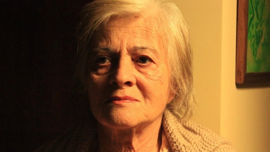 A foto mostra a atriz Araci Esteves, uma mulher idosa de pele clara com cabelos brancos e curtos, olhando diretamente para a câmera. Seu rosto, marcado por rugas, exibe uma expressão séria, quase melancólica. Ela veste um casaco de lã claro, de tricô. A iluminação lateral destaca as texturas de sua pele e do casaco, criando um forte contraste entre a luz à direita e a escuridão à esquerda do rosto e do fundo da imagem. A composição da foto é um close-up que foca na intensidade do olhar e na expressão da atriz, capturando a profundidade de seu personagem ou a emoção do momento. Um quadro com detalhes verdes é visível no canto superior direito.