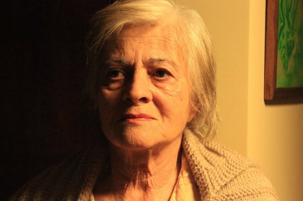 A foto mostra a atriz Araci Esteves, uma mulher idosa de pele clara com cabelos brancos e curtos, olhando diretamente para a câmera. Seu rosto, marcado por rugas, exibe uma expressão séria, quase melancólica. Ela veste um casaco de lã claro, de tricô. A iluminação lateral destaca as texturas de sua pele e do casaco, criando um forte contraste entre a luz à direita e a escuridão à esquerda do rosto e do fundo da imagem. A composição da foto é um close-up que foca na intensidade do olhar e na expressão da atriz, capturando a profundidade de seu personagem ou a emoção do momento. Um quadro com detalhes verdes é visível no canto superior direito.