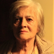 A foto mostra a atriz Araci Esteves, uma mulher idosa de pele clara com cabelos brancos e curtos, olhando diretamente para a câmera. Seu rosto, marcado por rugas, exibe uma expressão séria, quase melancólica. Ela veste um casaco de lã claro, de tricô. A iluminação lateral destaca as texturas de sua pele e do casaco, criando um forte contraste entre a luz à direita e a escuridão à esquerda do rosto e do fundo da imagem. A composição da foto é um close-up que foca na intensidade do olhar e na expressão da atriz, capturando a profundidade de seu personagem ou a emoção do momento. Um quadro com detalhes verdes é visível no canto superior direito.