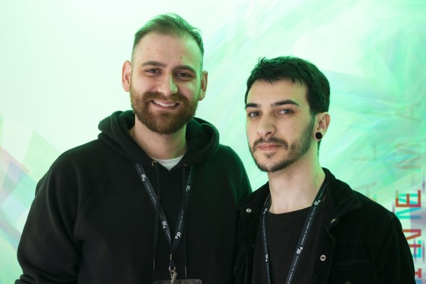 A foto mostra os artistas Márcio Picoli e Victor Di Marco, dois homens jovens, sorrindo e posando juntos. Márcio, à esquerda, tem barba cheia, cabelos curtos e castanhos claros. Ele veste um moletom preto com capuz. Victor, à direita, possui barba rala, cabelos escuros e curtos, e usa um casaco preto sobre uma camiseta escura. Ambos usam cordões de identificação de evento no pescoço. O fundo da foto é uma parede clara com uma iluminação colorida em tons de verde, criando um efeito suave e desfocado que destaca as duas figuras em primeiro plano.