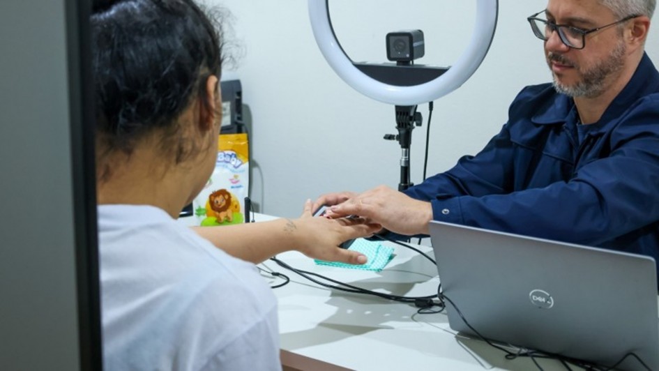 Uma foto em plano médio captura uma cena interna onde um homem e uma mulher estão sentados em uma mesa branca. O homem, à direita, usa óculos e uma camisa escura de manga comprida. Ele está segurando a mão direita da mulher, que está apoiada em um pequeno objeto retangular azul claro sobre a mesa. A mulher, vista por trás, veste uma camiseta branca e tem cabelo escuro preso. Na mesa, há um laptop cinza (marca Dell visível), uma luminária circular branca com uma câmera montada no topo e uma bolsa amarela com uma ilustração de leão. O fundo é uma parede branca lisa.