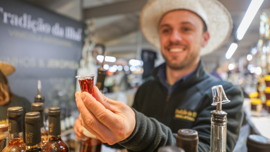 Uma foto em close-up mostra um homem sorrindo, usando um chapéu de palha de cor clara, segurando um pequeno copo de shot cheio de um líquido marrom-avermelhado. Ele está vestindo uma jaqueta escura e texturizada. Em primeiro plano, fora de foco, há várias garrafas, algumas com rótulos escuros e outras de vidro transparente. Uma garrafa à extrema direita tem um bico dosador decorativo. No fundo embaçado, há uma placa com um texto que parece dizer "Tradição do [ilegível]". O cenário parece ser um mercado interno ou uma banca, com iluminação superior brilhante visível.