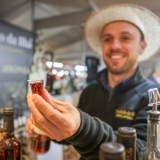 Uma foto em close-up mostra um homem sorrindo, usando um chapéu de palha de cor clara, segurando um pequeno copo de shot cheio de um líquido marrom-avermelhado. Ele está vestindo uma jaqueta escura e texturizada. Em primeiro plano, fora de foco, há várias garrafas, algumas com rótulos escuros e outras de vidro transparente. Uma garrafa à extrema direita tem um bico dosador decorativo. No fundo embaçado, há uma placa com um texto que parece dizer "Tradição do [ilegível]". O cenário parece ser um mercado interno ou uma banca, com iluminação superior brilhante visível.