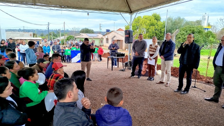A foto mostra um evento público ao ar livre, sob uma tenda de lona bege. Em primeiro plano e sentados no chão de terra, há um grupo de crianças e adultos olhando para um palco improvisado à frente. No centro da imagem, em pé, o Secretário de Habitação e Regularização Fundiária, Carlos Gomes, um homem de pele clara com óculos e camisa escura, discursa segurando um microfone. Ao lado dele, outros homens e uma mulher estão em pé, alguns com microfones e um deles ao lado de um banner com uma imagem de uma pessoa. Ao fundo, casas simples e áreas verdes completam o cenário.