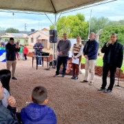A foto mostra um evento público ao ar livre, sob uma tenda de lona bege. Em primeiro plano e sentados no chão de terra, há um grupo de crianças e adultos olhando para um palco improvisado à frente. No centro da imagem, em pé, o Secretário de Habitação e Regularização Fundiária, Carlos Gomes, um homem de pele clara com óculos e camisa escura, discursa segurando um microfone. Ao lado dele, outros homens e uma mulher estão em pé, alguns com microfones e um deles ao lado de um banner com uma imagem de uma pessoa. Ao fundo, casas simples e áreas verdes completam o cenário.