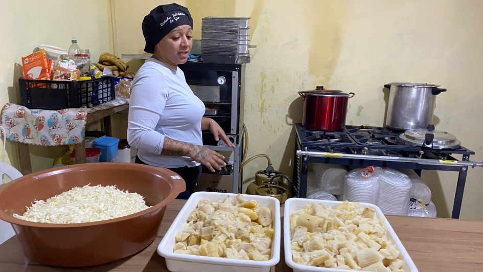 A foto mostra uma mulher jovem, com cabelos presos e vestindo uma blusa branca de manga comprida e um boné preto. Ela está em pé em uma cozinha e gesticula enquanto olha para a esquerda, aparentemente conversando com alguém fora da foto. Sobre uma mesa de madeira, há três grandes travessas: uma bacia marrom com um alimento ralado branco, e duas travessas brancas com pedaços de frango e queijo já misturados, que parecem prontos para o preparo. Ao fundo, uma geladeira, um fogão industrial com panelas, e diversos utensílios e ingredientes de cozinha compõem a cena, sugerindo a preparação de uma grande quantidade de alimentos.
