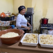 A foto mostra uma mulher jovem, com cabelos presos e vestindo uma blusa branca de manga comprida e um boné preto. Ela está em pé em uma cozinha e gesticula enquanto olha para a esquerda, aparentemente conversando com alguém fora da foto. Sobre uma mesa de madeira, há três grandes travessas: uma bacia marrom com um alimento ralado branco, e duas travessas brancas com pedaços de frango e queijo já misturados, que parecem prontos para o preparo. Ao fundo, uma geladeira, um fogão industrial com panelas, e diversos utensílios e ingredientes de cozinha compõem a cena, sugerindo a preparação de uma grande quantidade de alimentos.